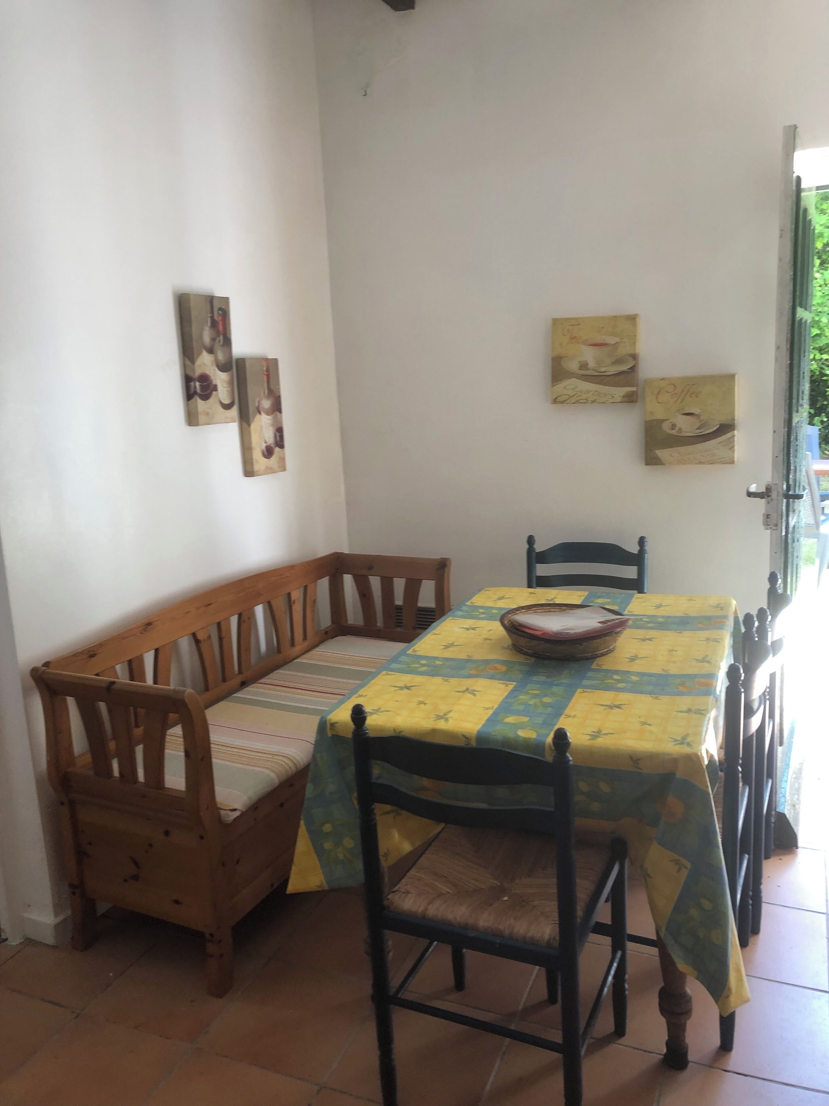 A dining area with a wooden bench and chairs, a table covered with a yellow tablecloth, and two wall-mounted paintings.