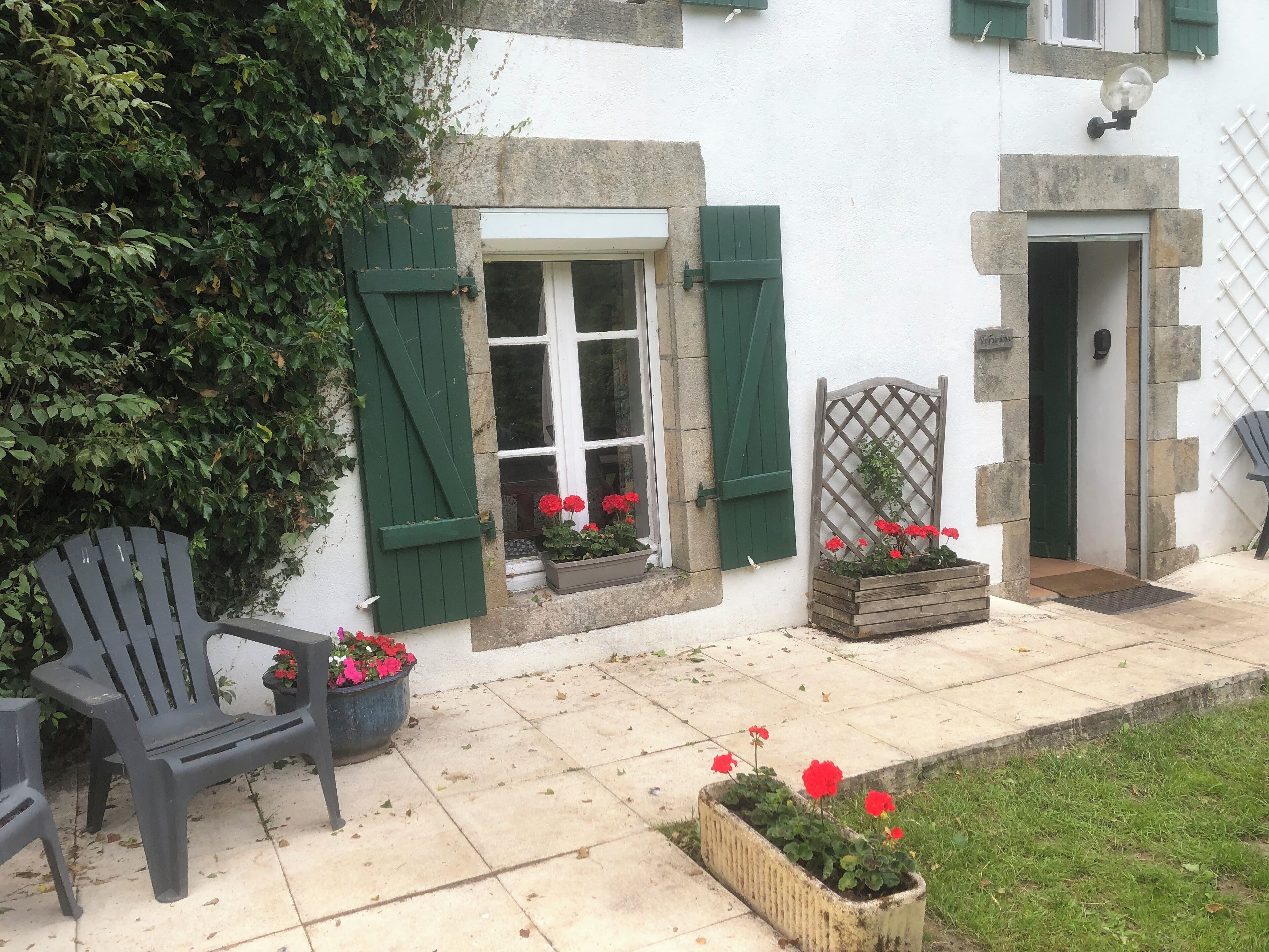 A quaint building with a white facade, green shutters, and a wooden bench. There are flower pots with red flowers on the steps and a wooden gate.