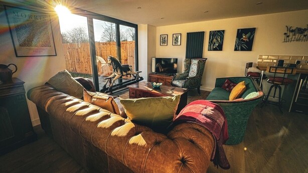 Un salon chaleureux avec un canapé en cuir, un fauteuil vert et une table basse en bois. Une grande fenêtre offre une vue sur une clôture en bois et une sculpture équestre.