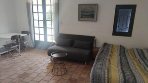 A room with a sofa, a small round table, a desk with chairs, and a bed with a patterned blanket. - Studio in the countryside (Le Thor)