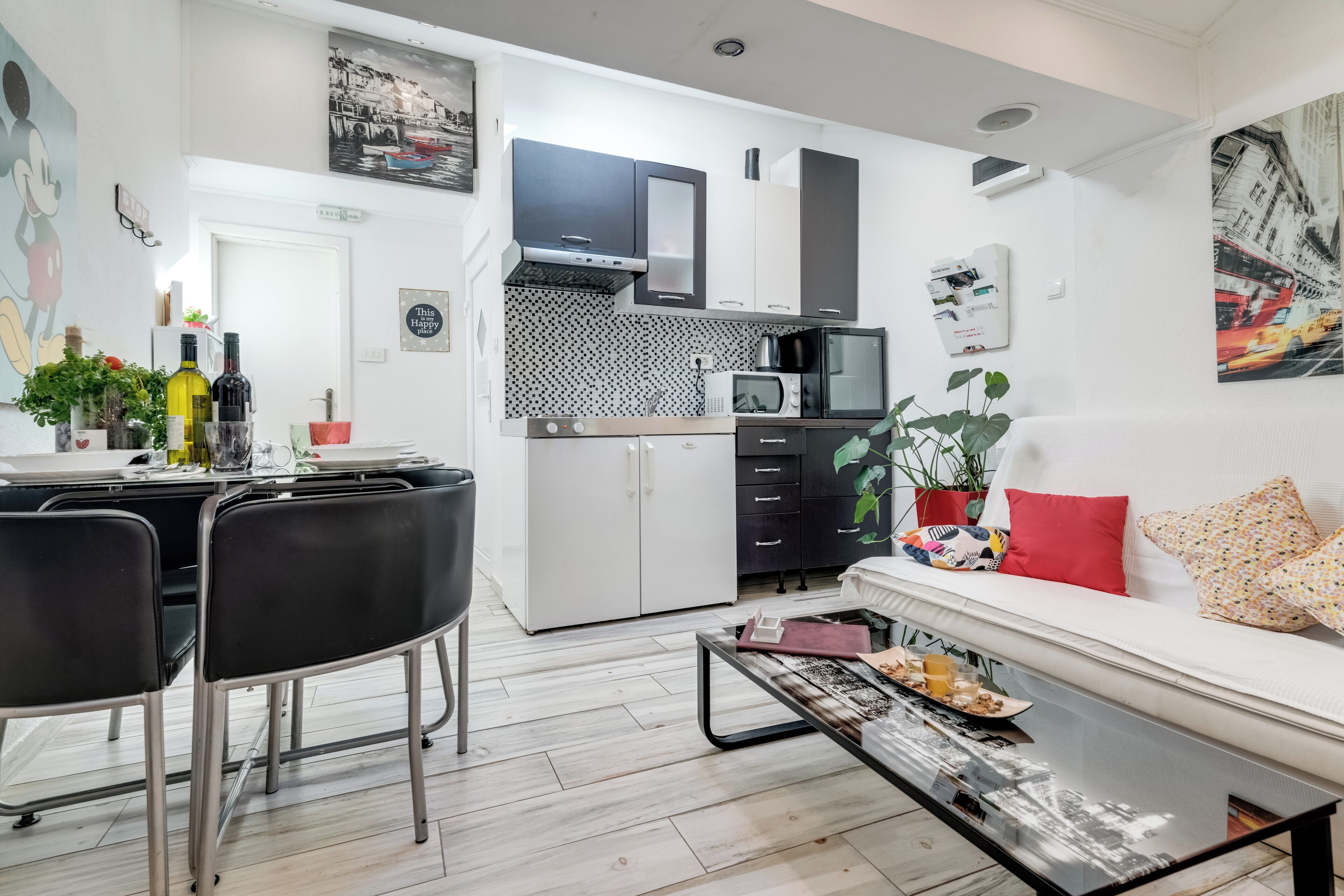 Een moderne keuken met een magnetron, fornuis en kastjes. Er is een zithoek met een tafel en stoelen. Een bank met een rood kussen bevindt zich in de woonkamer.