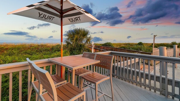 Une terrasse en bois avec une table et des chaises, un parasol à rayures et une vue sur la plage et le ciel.