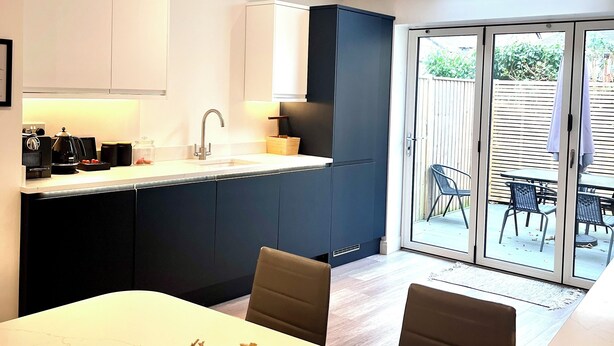 A modern kitchen with a white countertop, dark cabinets, and a dining area with a white table and chairs. - Mode Cottage (Lytham St. Anne's)