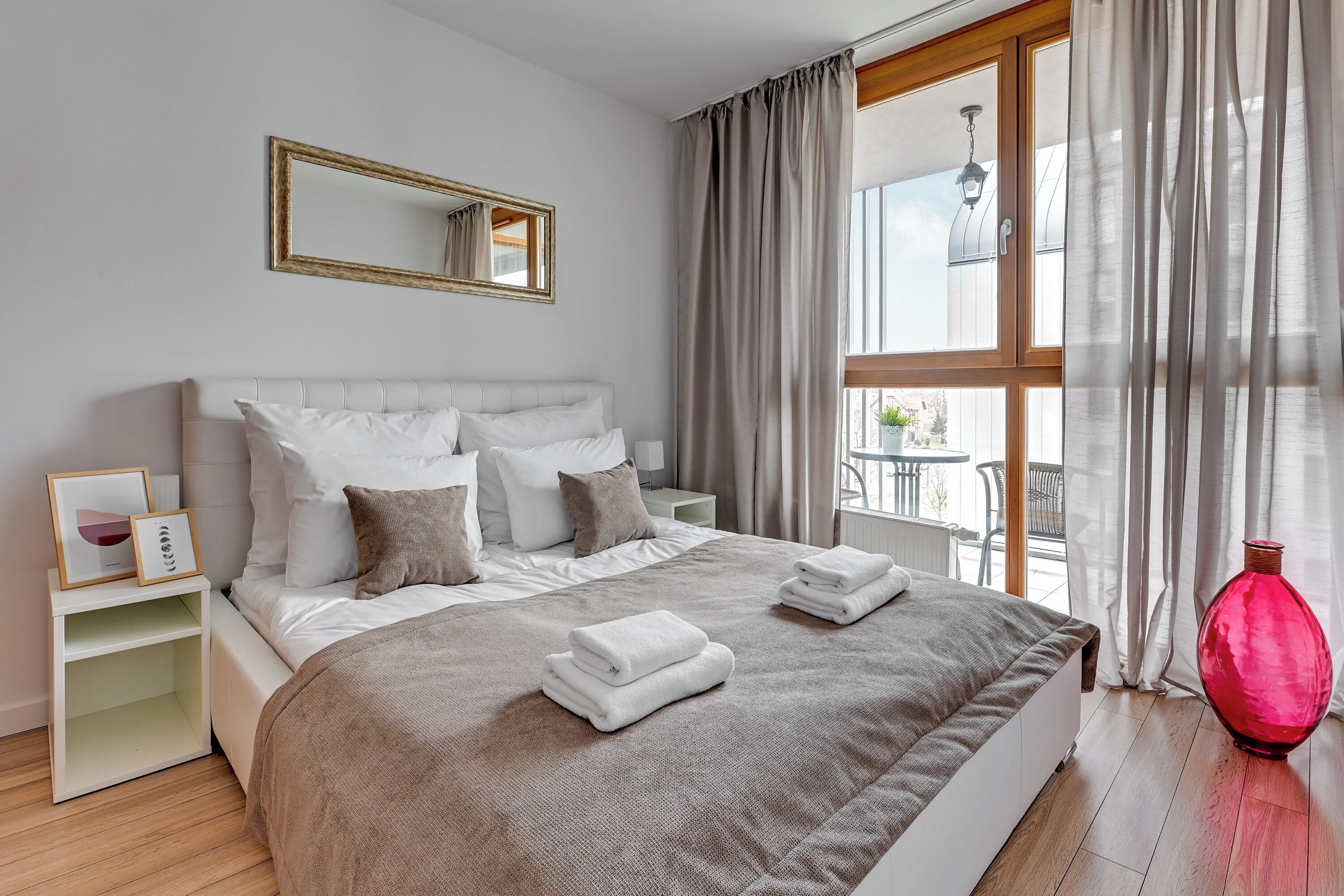 A neatly made bed with white bedding and a brown throw, a bedside table with a lamp, a large mirror, and a view of the outdoors through a window with curtains.
