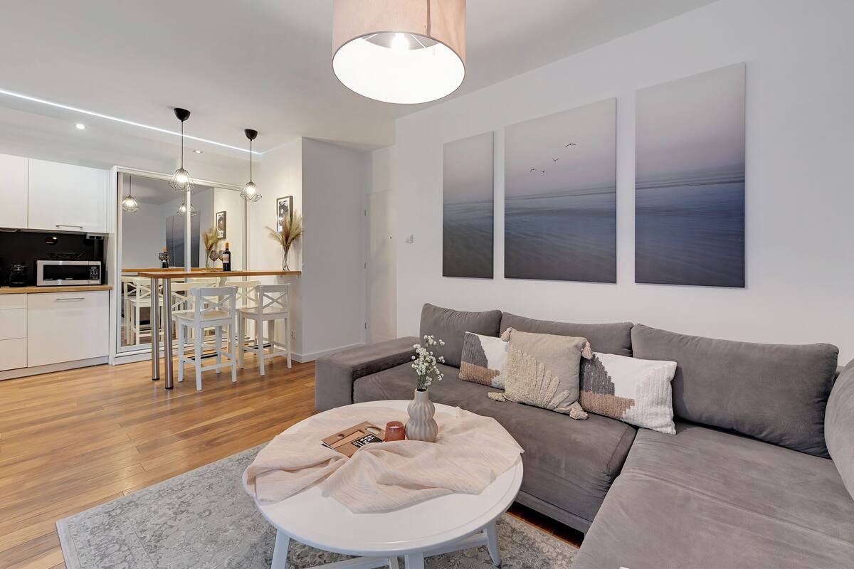 A modern living room with a grey sofa, a round coffee table with a vase and candles, and a dining area with white chairs and a wooden table.