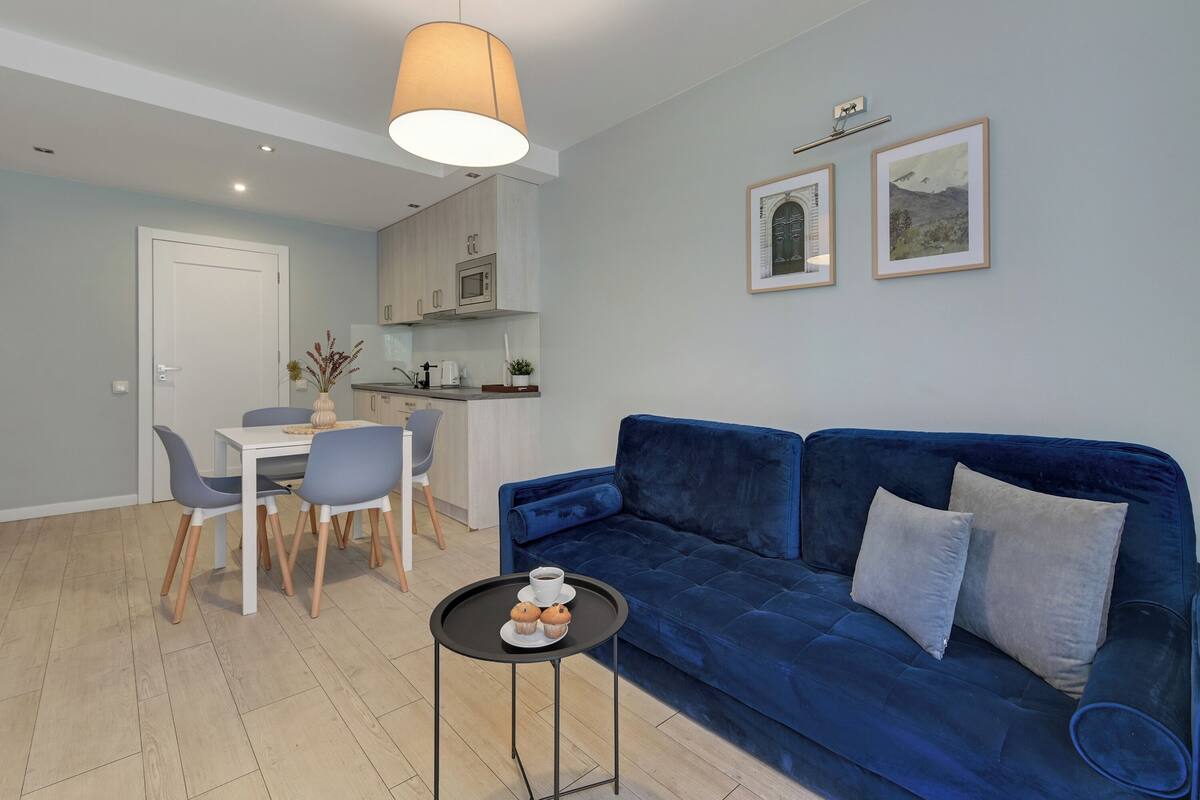 A modern living room with a blue sofa, a dining table with chairs, a small coffee table, and a kitchen area in the background.