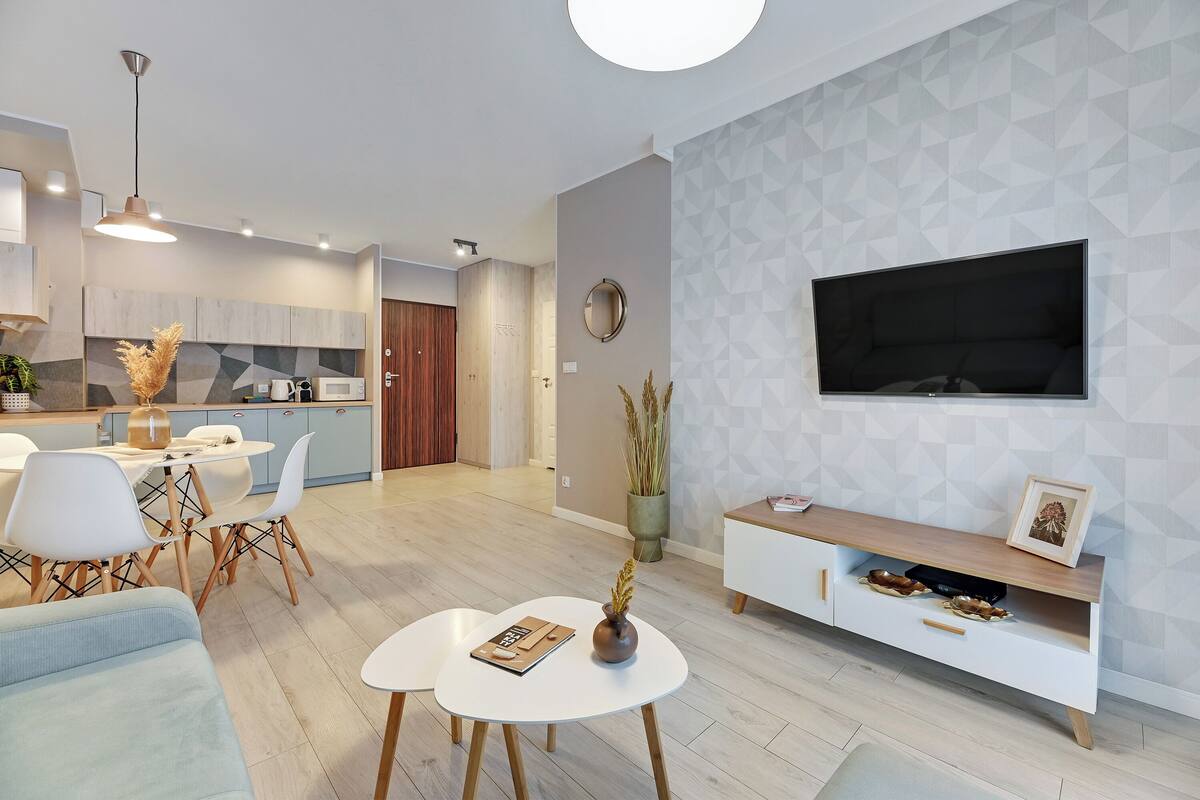 A modern living room with a white sofa, a coffee table, and a TV mounted on the wall. There is a kitchen area in the background with wooden cabinets and a dining table.