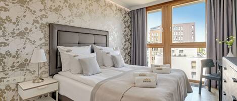 A modern hotel room with a large bed, a nightstand, a lamp, a dresser, and a view of the city through large windows.
