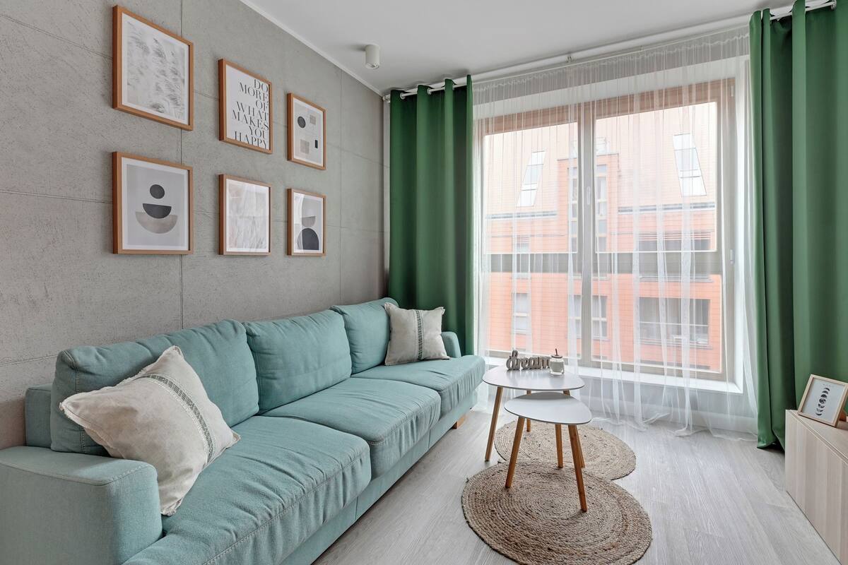 A modern living room with a teal sofa, a small round table, and a large window with green curtains.
