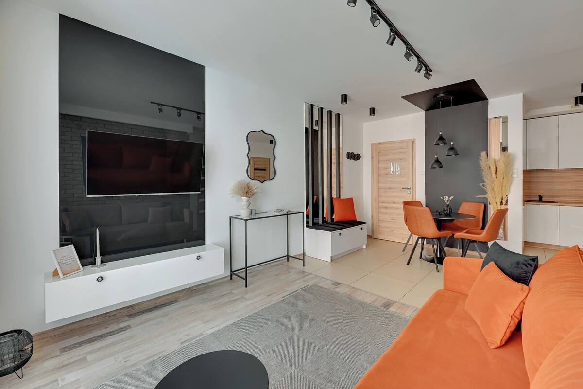 A modern living room with a large TV, a dining area with orange chairs, and a kitchen with white cabinets.