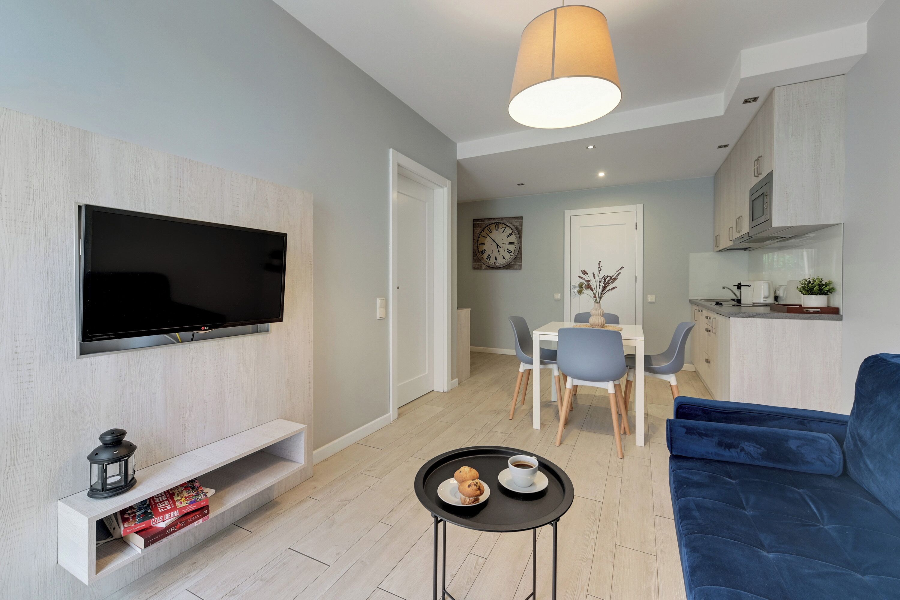 A modern living room with a flat-screen TV, a blue sofa, a small coffee table, and a dining area with a white table and chairs.