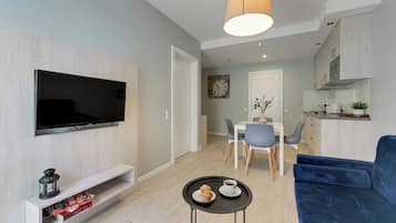 A modern living room with a flat-screen TV, a blue sofa, a small coffee table, and a dining area with a white table and chairs.