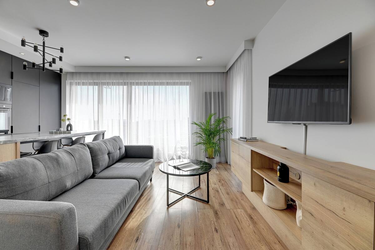 A modern living room with a grey sectional sofa, a glass coffee table, a wooden TV stand, and a large flat-screen TV mounted on the wall.