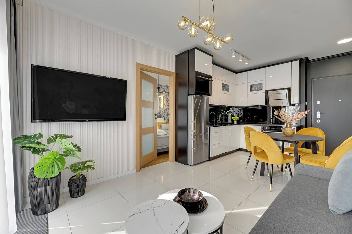 A modern kitchen with a built-in oven, a dining area with yellow chairs, and a flat-screen TV mounted on the wall.