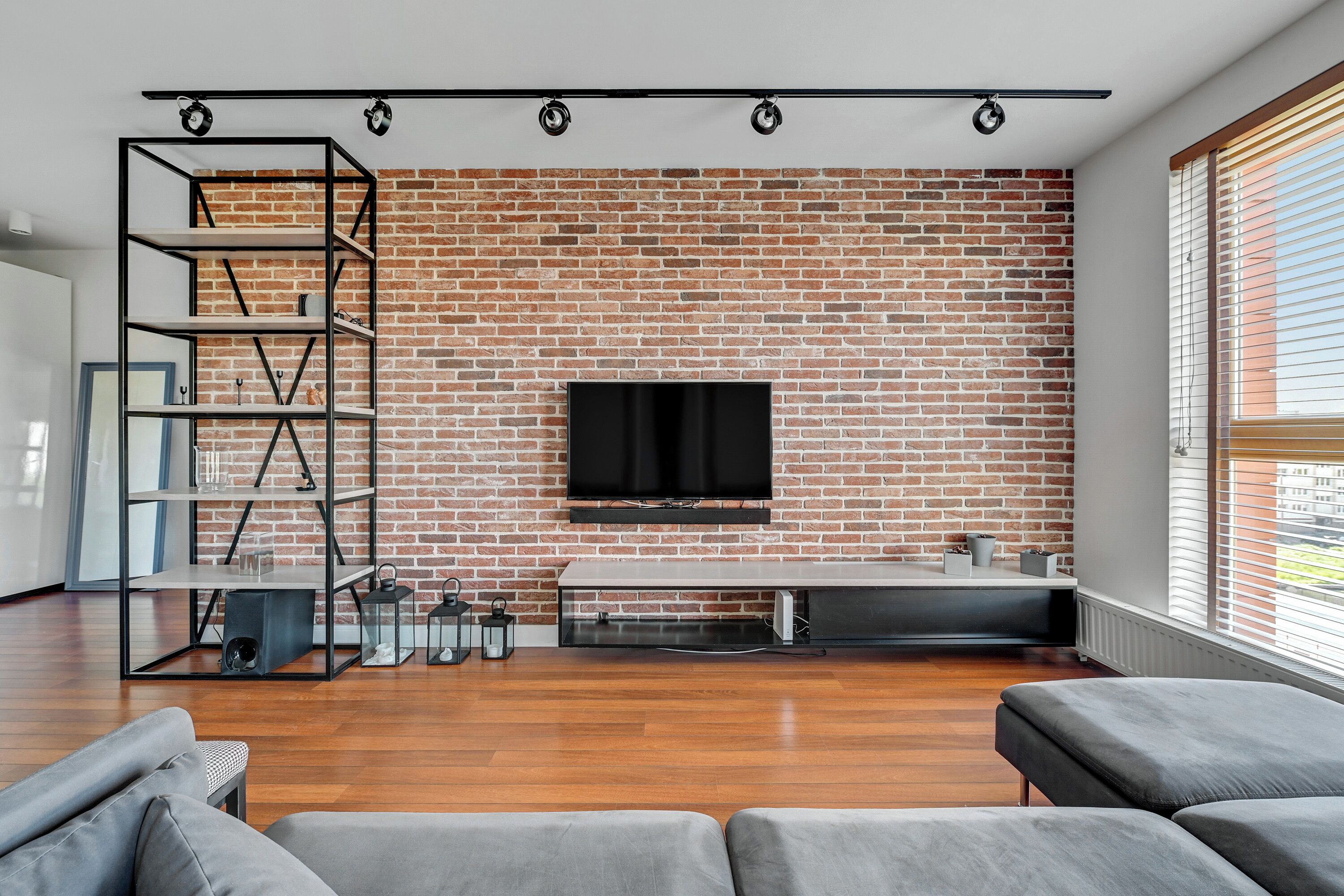 A modern living room with a brick wall, a flat-screen TV, a wooden floor, and grey sofas.