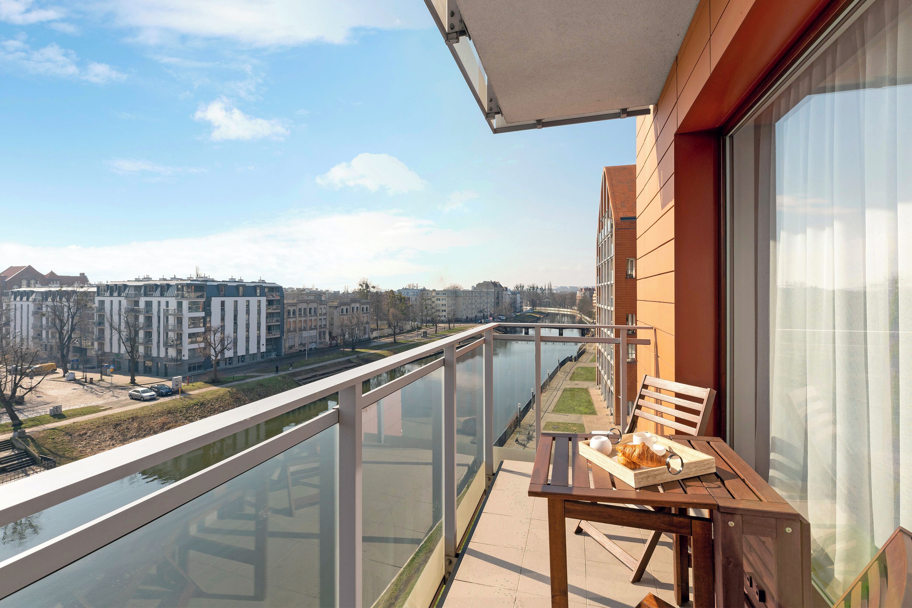 A balcony with a view of a river and buildings, a table with a chair, and a window with curtains.