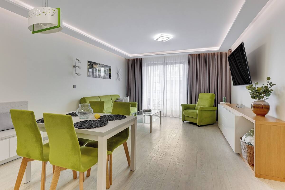 A modern living room with a dining area, green chairs, a white table, and a flat-screen TV mounted on the wall.