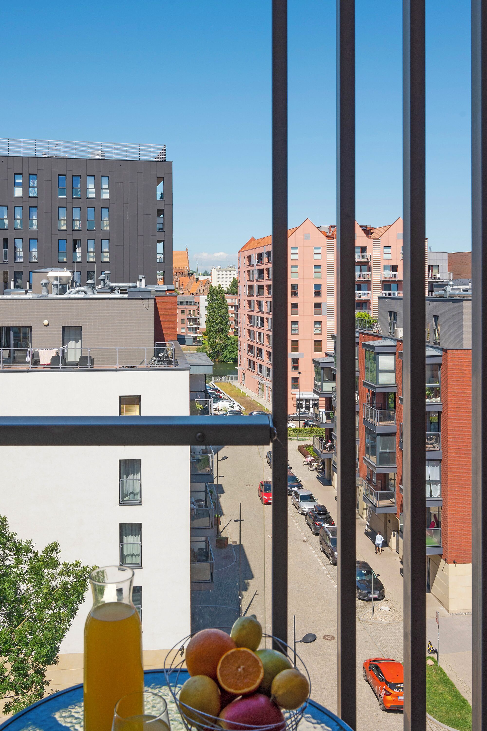 A balcony with a beverage and fruit basket, overlooking a city street with buildings and parked cars.