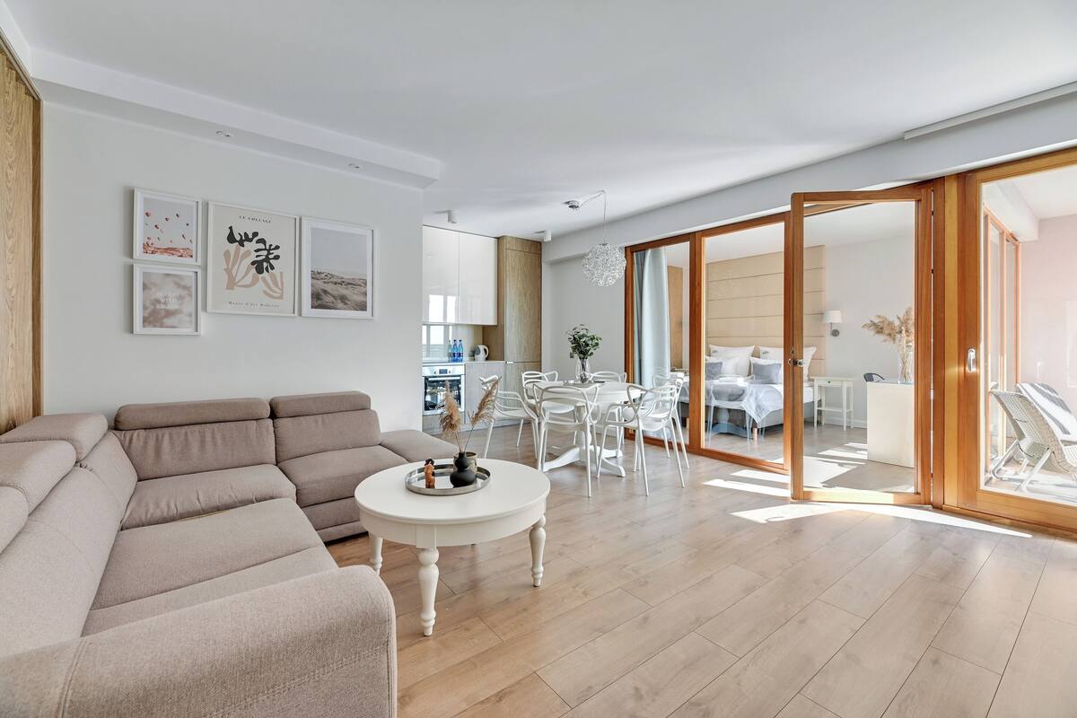 A modern living room with a beige sectional sofa, a white round coffee table, and a dining area with white chairs and a glass-top table.