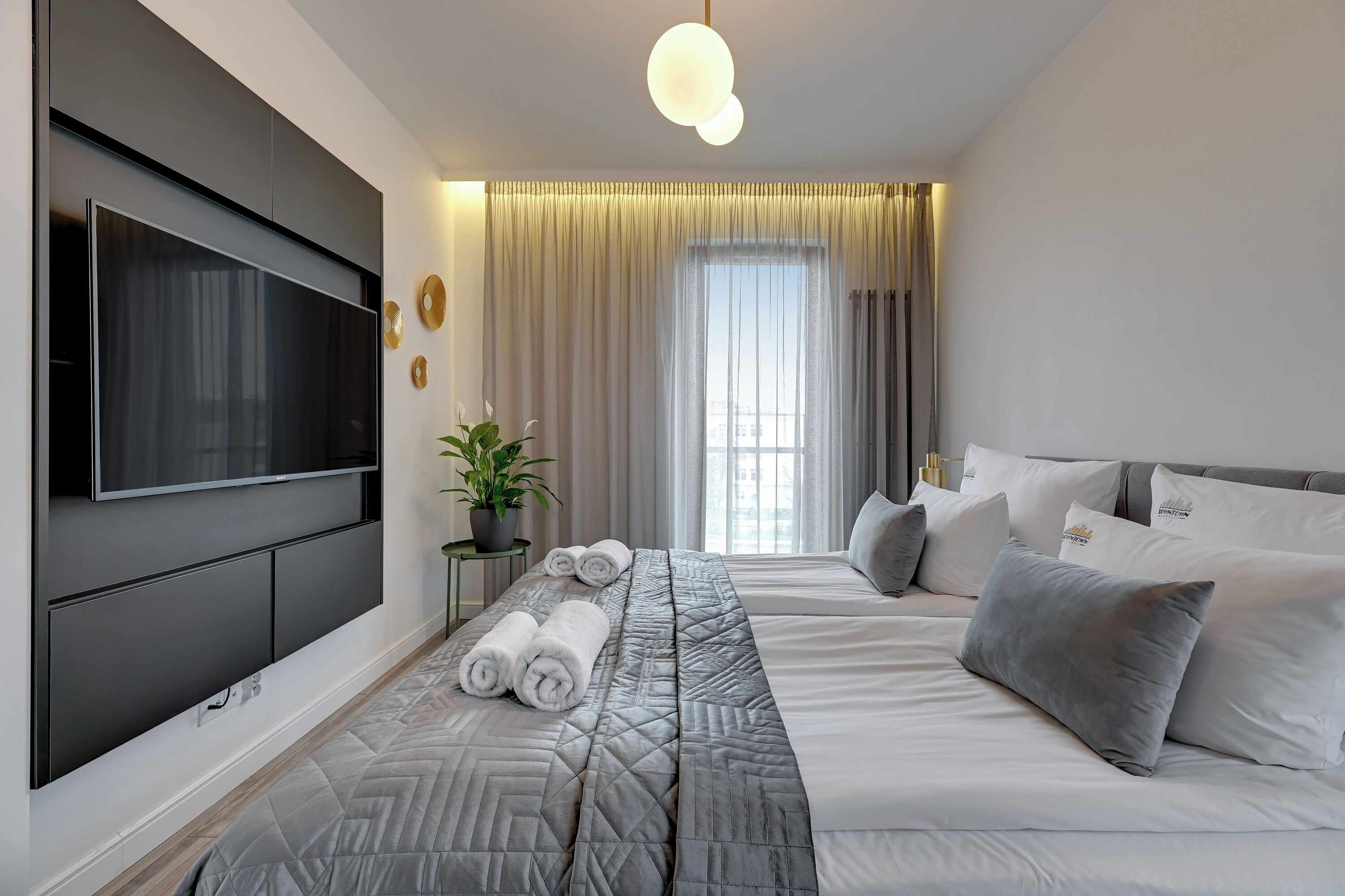 Une chambre d’hôtel moderne dotée d’un grand téléviseur à écran plat fixé au mur, d’un lit avec une literie grise et d’une plante en pot posée sur une table d’appoint.