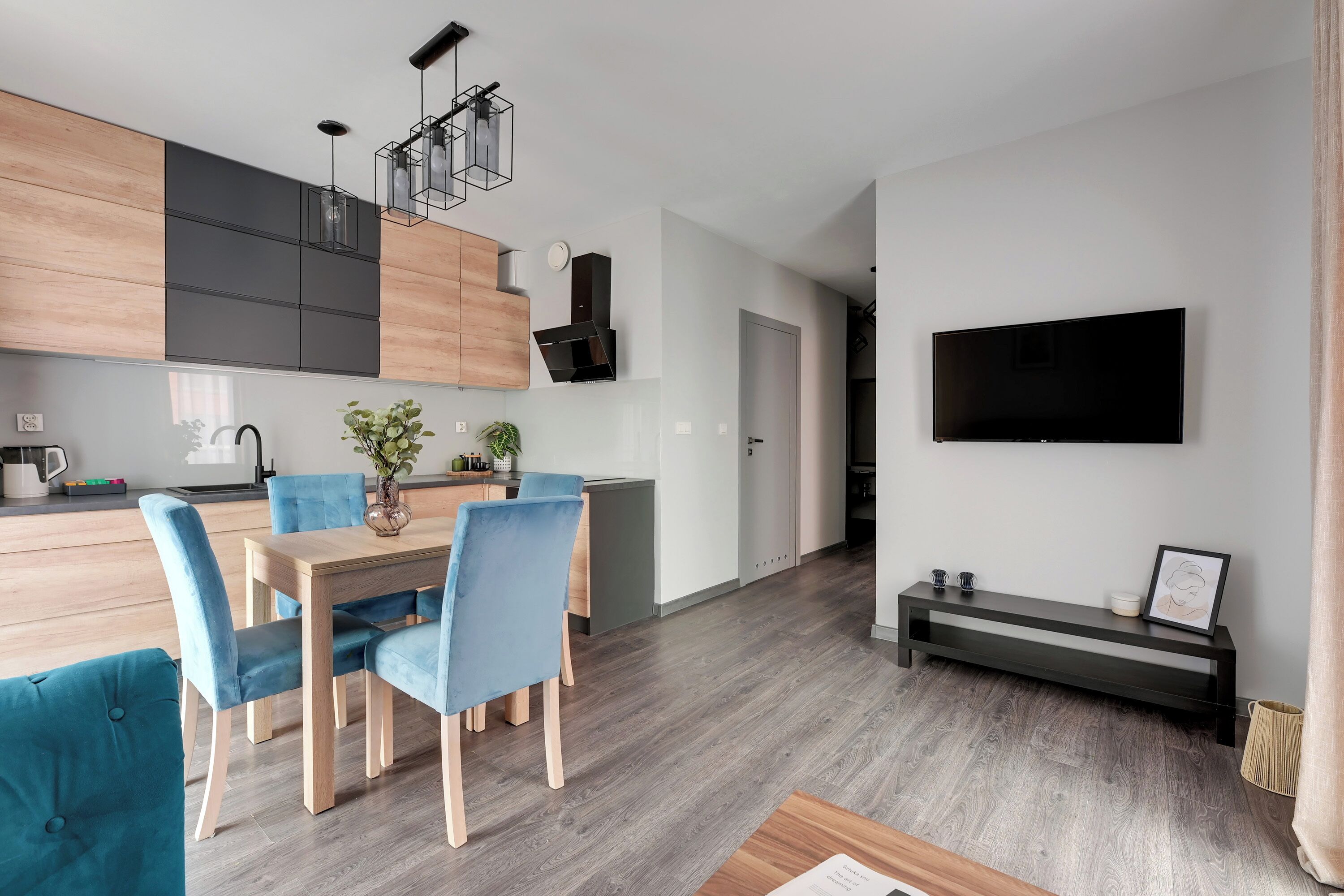 A modern kitchen with wooden cabinets, a dining area with blue chairs, and a flat-screen TV mounted on the wall.