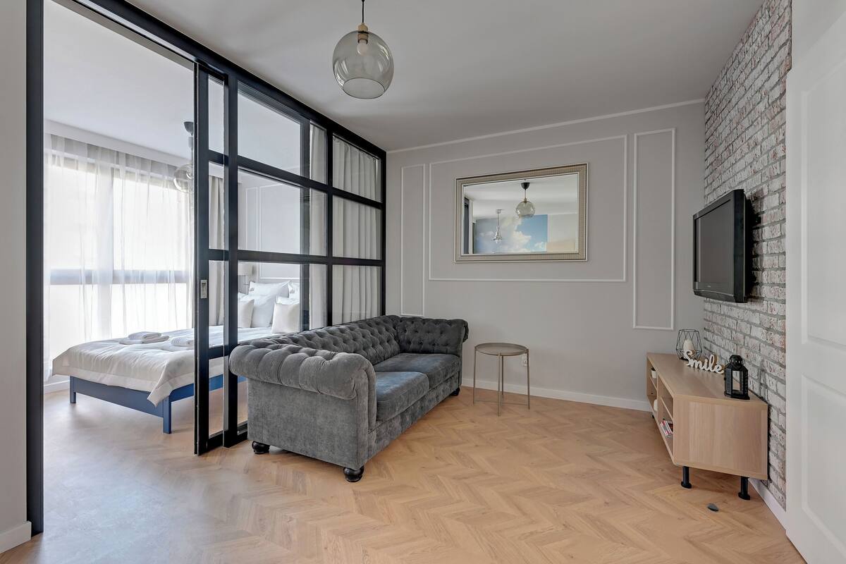 A modern bedroom with a grey sectional sofa, a bed with white linens, a small round table, and a TV mounted on a brick wall.