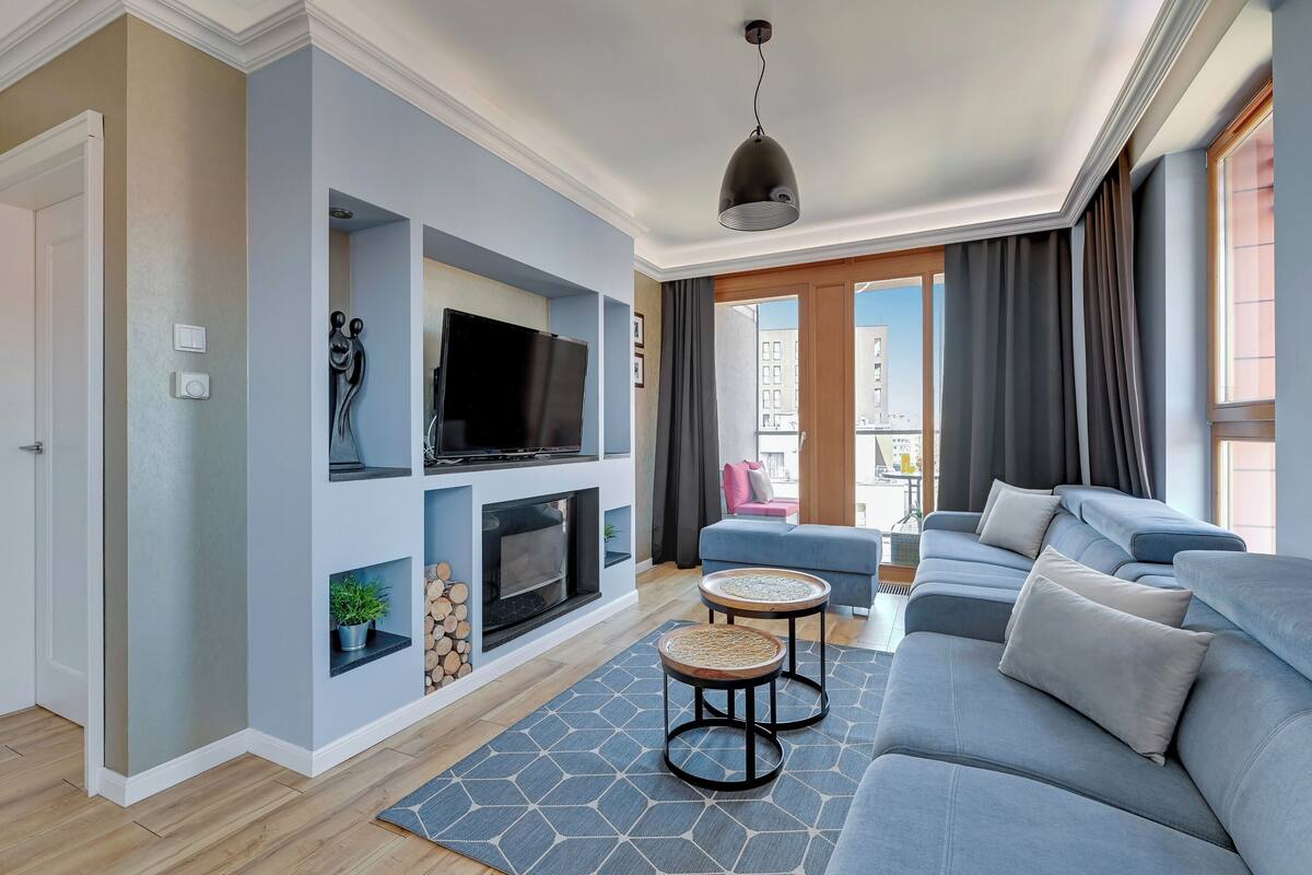 A modern living room with a grey sofa, a flat-screen TV, a fireplace, and a balcony with a view of buildings.