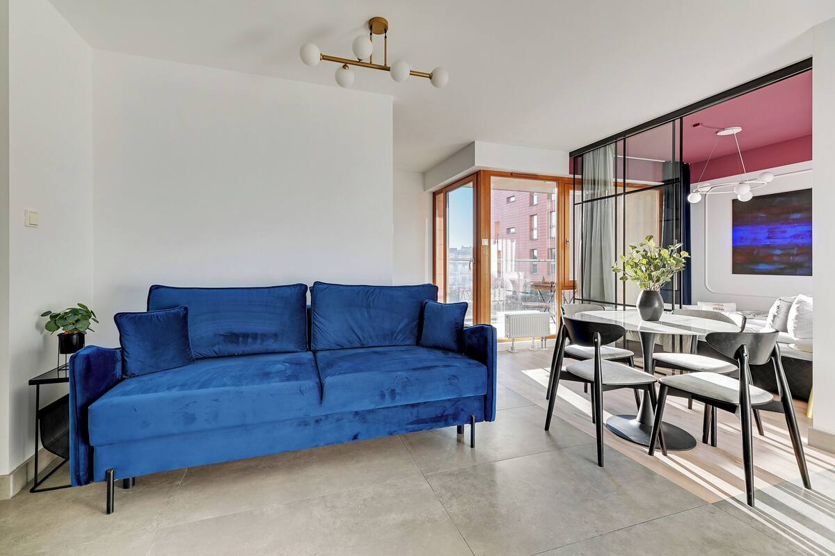 A modern living room with a blue sofa, a dining table with chairs, a potted plant, and a large window offering a view of buildings.