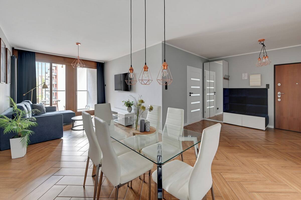 A modern living room with a glass dining table, white chairs, and a sofa. There is a TV mounted on the wall, a potted plant, and a wooden floor.