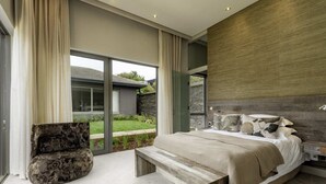 A modern bedroom with a large bed, a bench, and a view of a garden through large windows. - Cliff Break (Knysna)