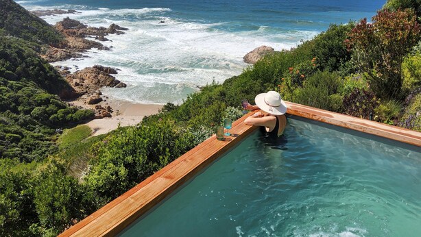 A person relaxing in a wooden infinity pool overlooking a beach and ocean. - Cliff Break (Knysna)