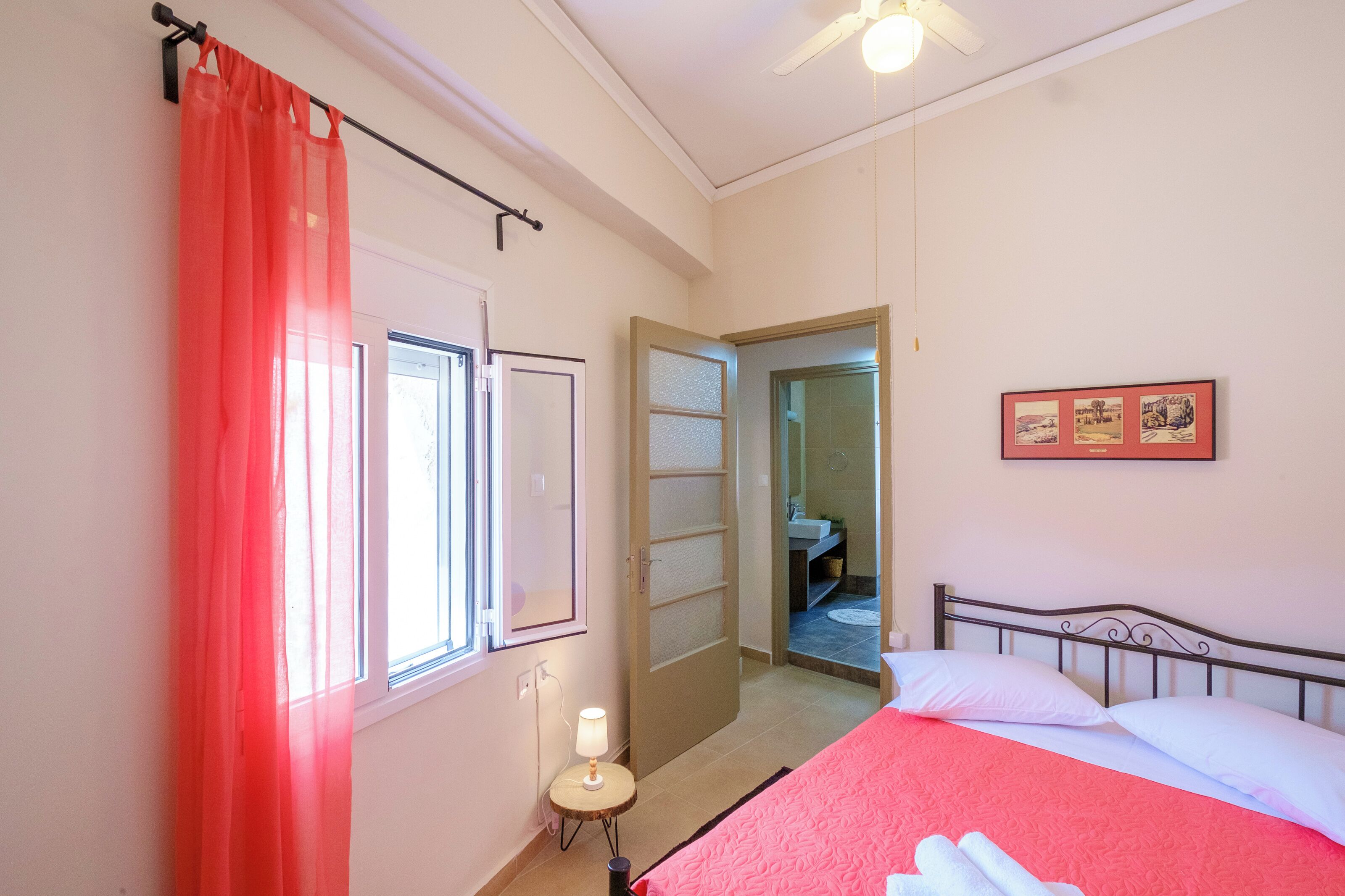 Een slaapkamer met een bed, een raam met gordijn, een nachtkastje met lamp en een deur naar een andere kamer.