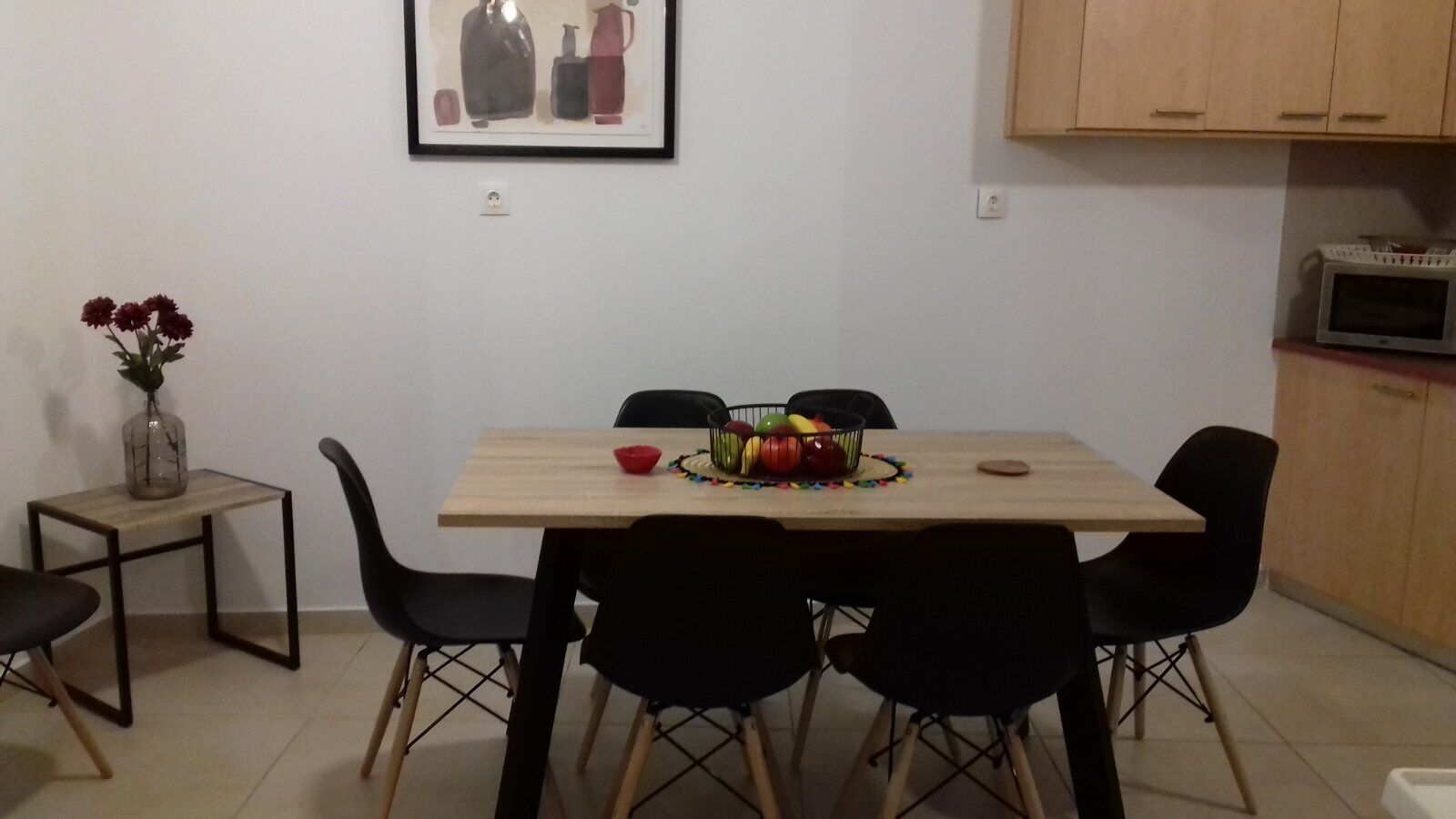 A dining area with a wooden table, black chairs, a small side table with a vase of flowers, and a framed picture on the wall.