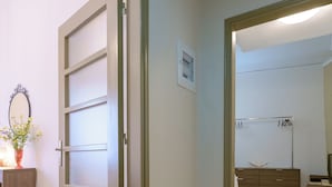 A room with a wooden floor, a white door, a mirror, a small table with a vase of flowers, and a wardrobe with a hanging rod.