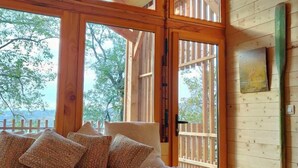 A wooden cabin with a large window, a sofa with cushions, a small table, and a wall-mounted light fixture. - La Cabane De La Saône, Lyon Country House (Caluire et Cuire)