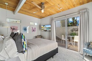 Une chambre avec un lit, un ventilateur de plafond, des œuvres d’art accrochées aux murs et une porte-fenêtre coulissante donnant sur un espace extérieur équipé d’une table et de chaises.