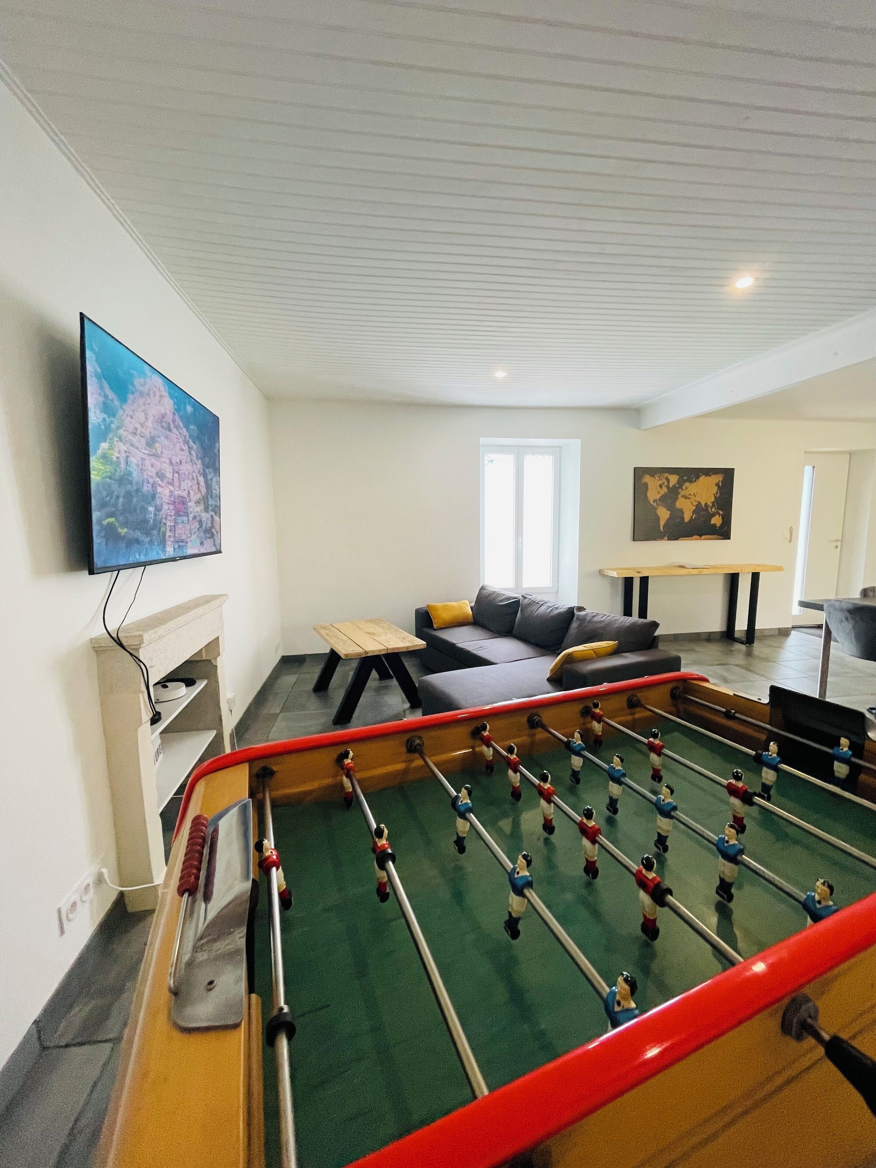 Um quarto com mesa de pebolim, televisão na parede, sofá e uma mesinha de cabeceira com abajur.