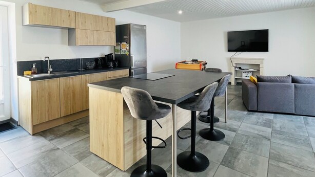 A modern kitchen with wooden cabinets, a central island with bar stools, a flat-screen TV, and a grey sofa in a living area. - House near the sea!  (Grues)