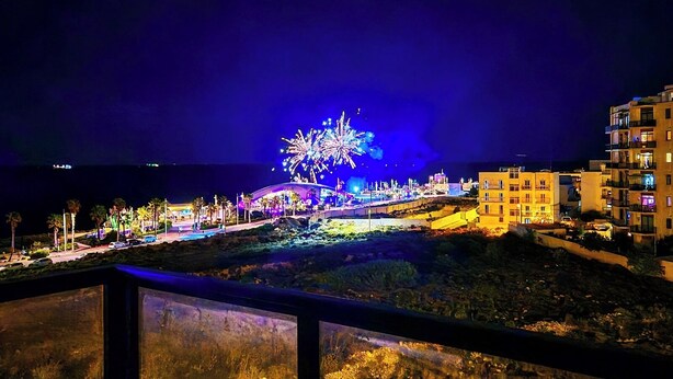 A nighttime view of a coastal area with a brightly lit fireworks display, buildings, and a road.