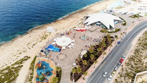 An aerial view of a coastal area with a large white building, a playground, and a road with vehicles.