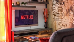 A living room with a flat-screen TV displaying 'NETFLIX,' a shelf with decorative items, a wooden coffee table, and a wall with a vintage newspaper design.