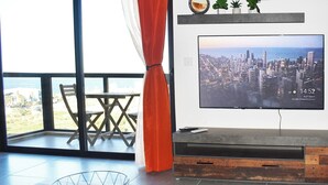 A modern living room with a large TV displaying a cityscape, a dining area with a table and chairs, and a view of the outdoors.