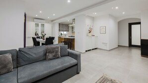A modern living room with a grey sofa, a dining area with a table and chairs, a kitchen with white cabinets, and a hallway leading to a door.