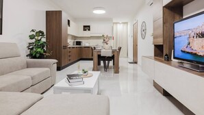 Un salon moderne avec un canapé, une table basse et un téléviseur. Au fond, on aperçoit une cuisine avec une table à manger et des chaises.