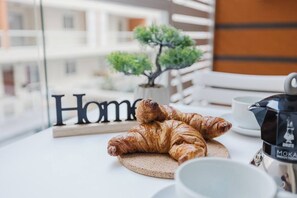 Un croissant sur une planche à découper ronde en bois, une cafetière et une tasse sur une table, avec vue sur un bâtiment et une enseigne portant le mot « Accueil ».