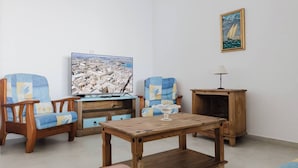 Ein Wohnzimmer mit Couchtisch aus Holz, zwei Sesseln, einem Fernseher, einer Lampe und einem gerahmten Bild an der Wand.
