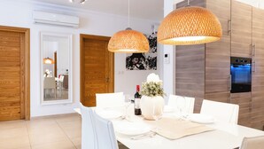 A modern dining area with a white table set for four, two pendant lights, and a kitchen in the background. - 102 Seafront 3Bed Sliema with pool by Homely (Tas-Sliema)