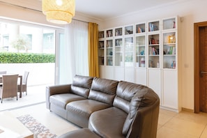 Un salon moderne avec un canapé en cuir, une porte vitrée donnant sur l’extérieur et une étagère blanche où sont disposés des livres et des objets décoratifs.