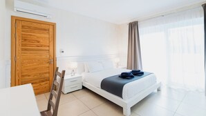 A hotel room with a wooden door, a white bed, a nightstand with a lamp, and a window with curtains. - 102 Seafront 3Bed Sliema with pool by Homely (Tas-Sliema)
