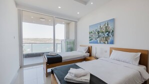 A modern hotel room with a large bed, two smaller beds, a balcony with a view of the sea, and a painting on the wall. - This apartment is a 2 bedroom(s), 1 bathrooms, located in Il-Mellieħa, null. (Il-Mellieħa)